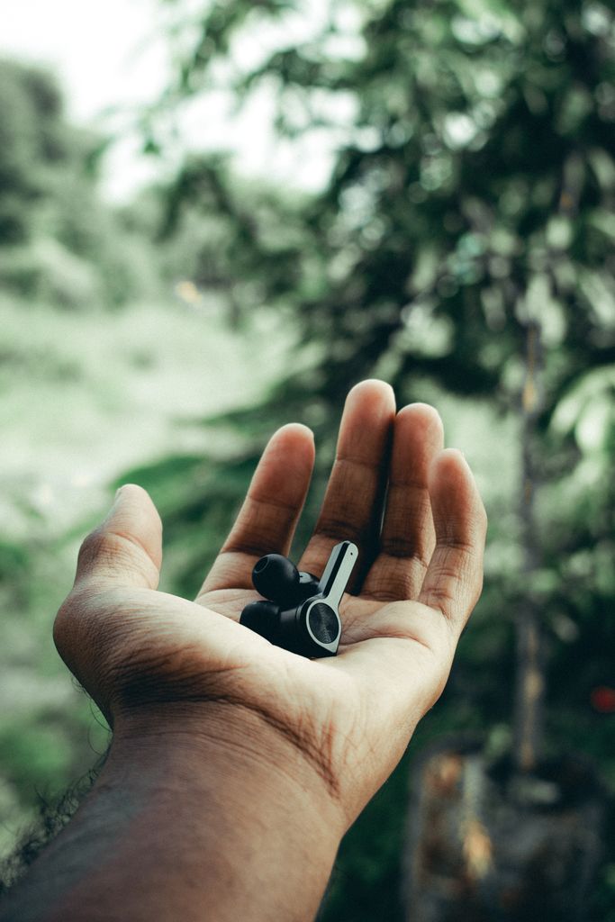 A hand holds black wireless earbuds against a lush outdoor background, highlighting modern technology.