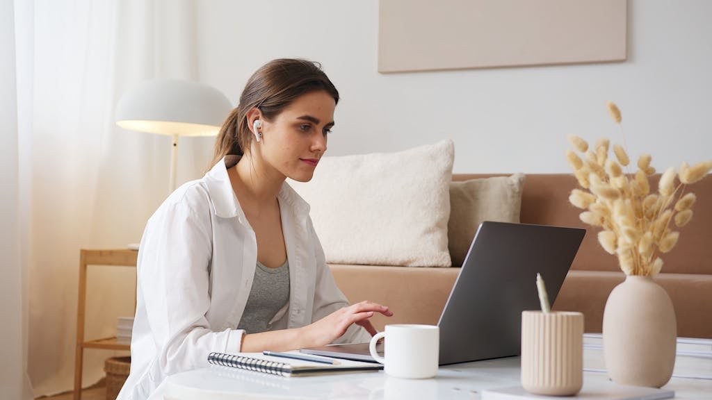Positive focused young female in casual clothes with wireless earbuds browsing netbook while listening to favorite music and sitting on floor in cozy apartment