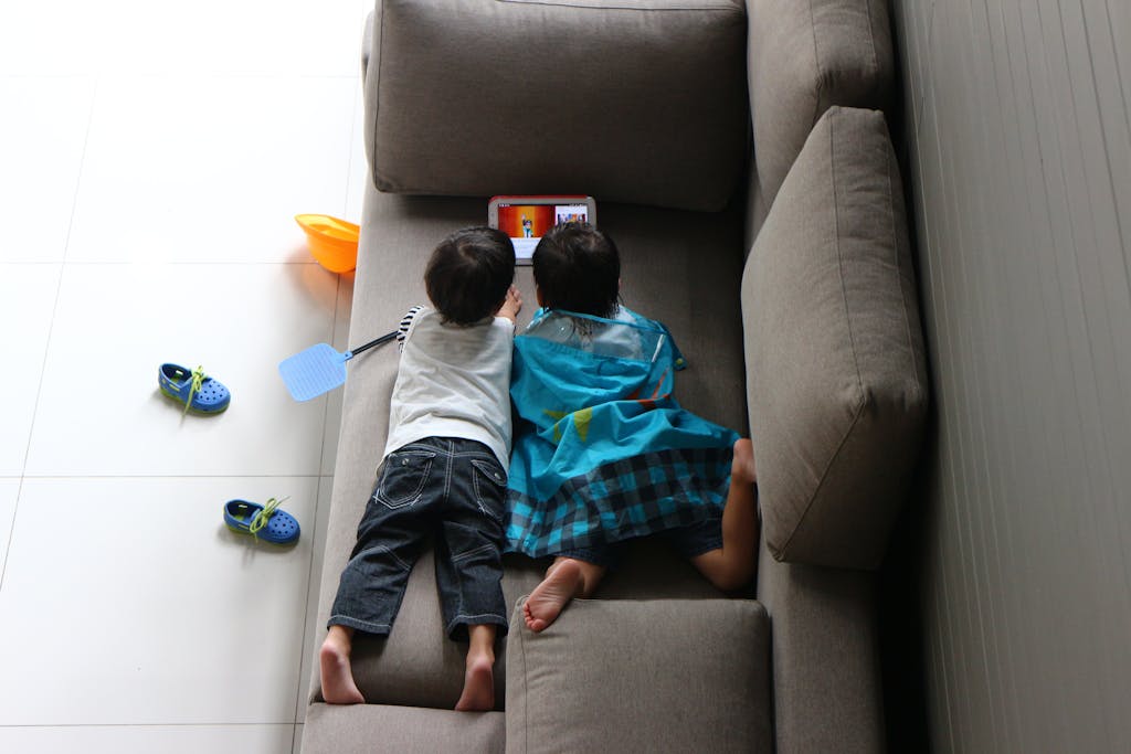 Two children lie on a sofa watching a tablet, enjoying indoor playtime.