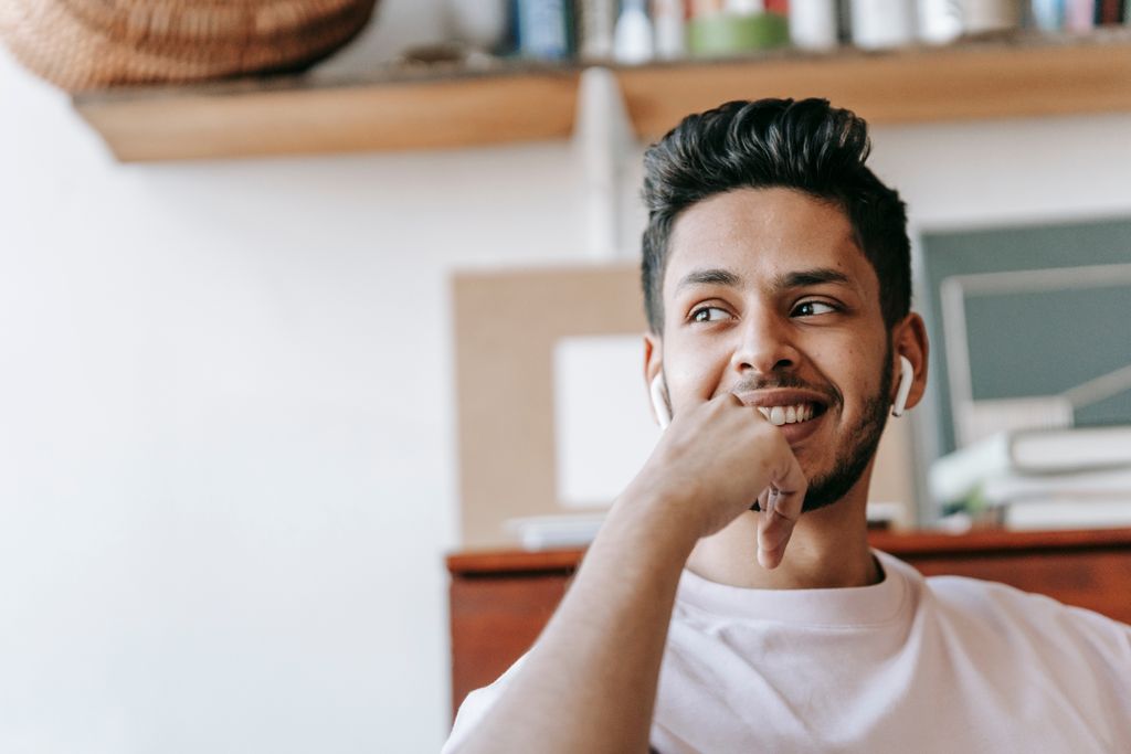 Cheerful young ethnic male listening to good music via wireless earbuds and looking away with smile while sitting in light living room and touching chin