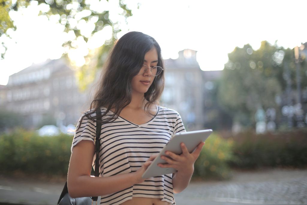 From below thoughtful young woman focusing on screen and interacting with digital tablet while standing on street against blurred trees and buildings in city